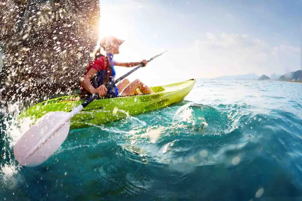 Young lady paddling hard the kayak with lots of splashes near the cliff at sunny day