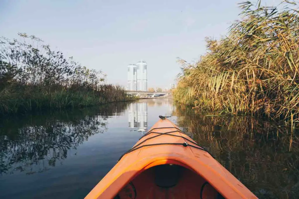 Red kayak nose rowing towards modern buildings through the forest on the waters of Dnipro river in Kyiv, Ukraine.