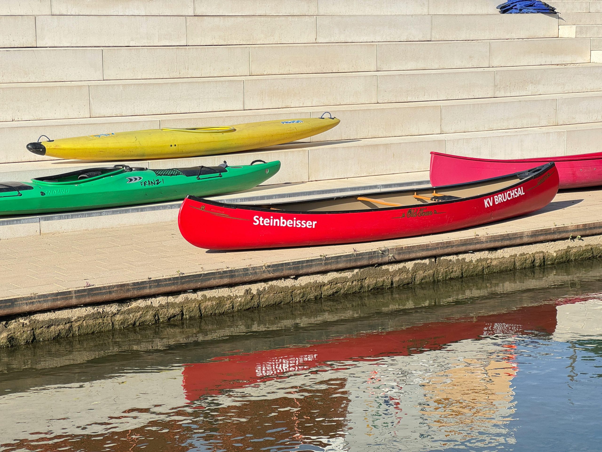 Colorful canoes rest on steps by the waterside in Bruchsal, Germany, with vivid reflections.