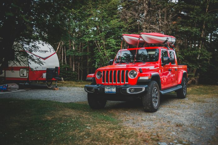 Red off-road truck with kayaks parked at a forest campsite with trailer nearby.