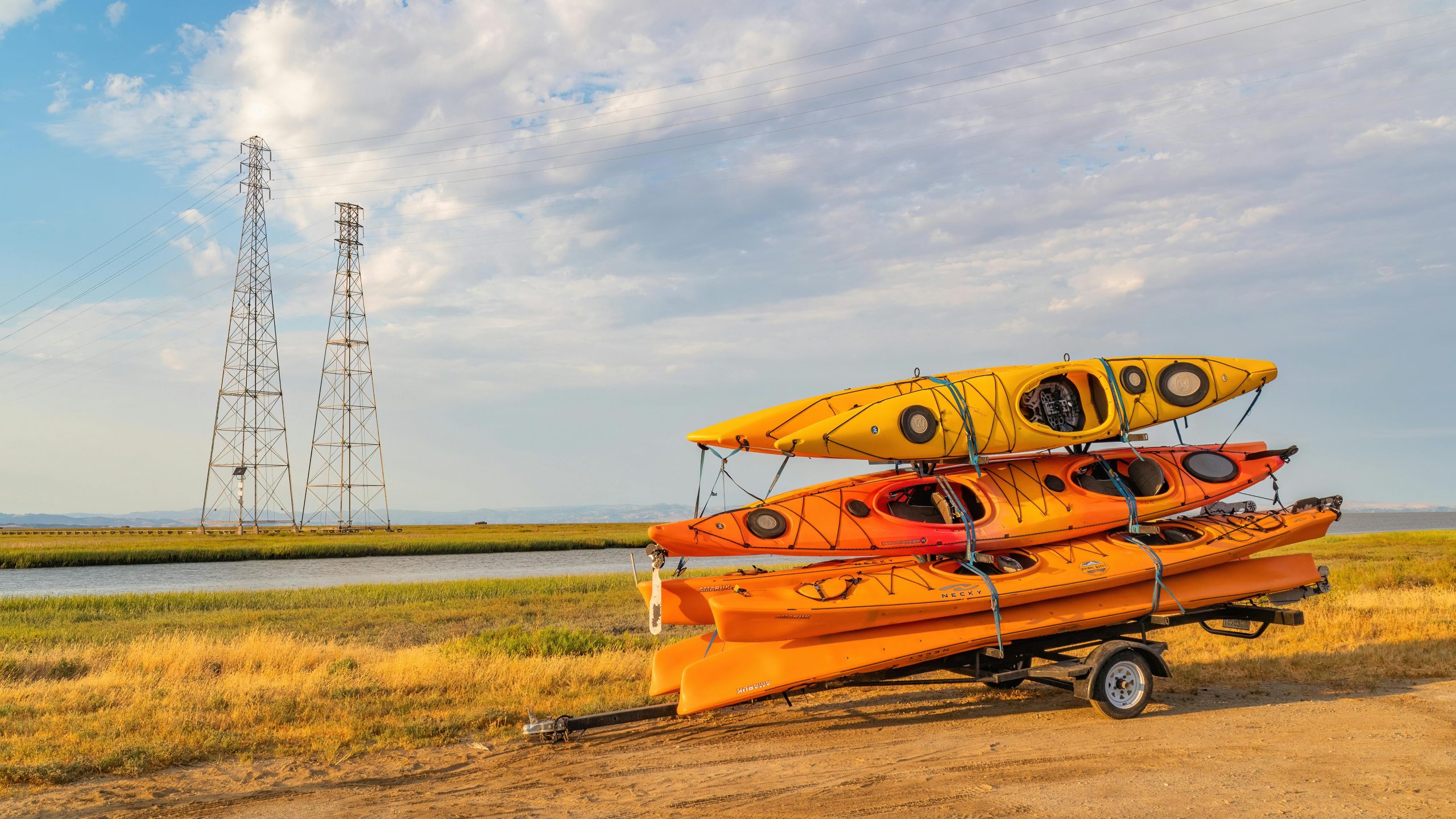 A stack of vibrant kayaks on a trailer by the water, under a blue sky.
