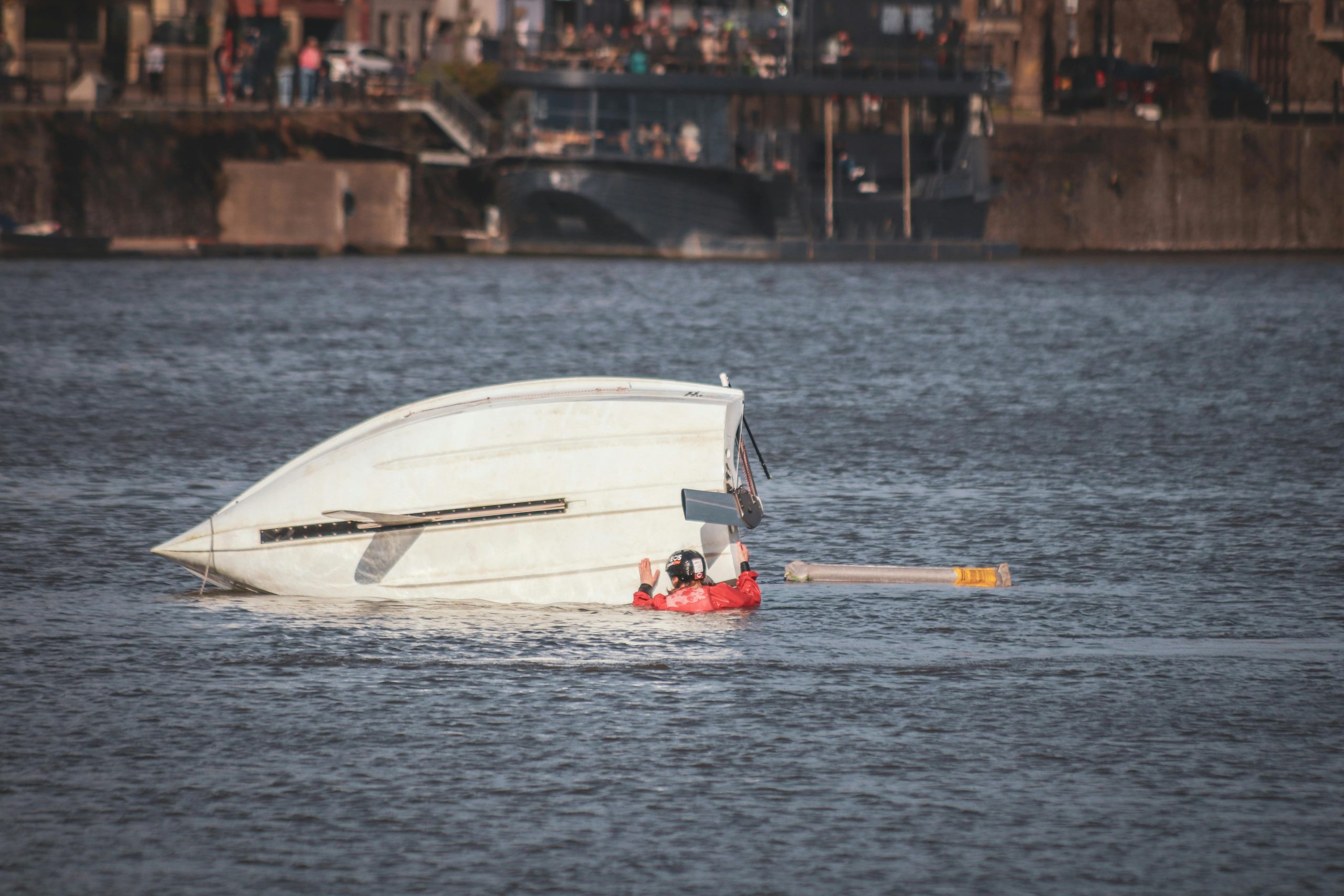A capsized boat near an urban waterfront with a person wearing a life jacket visible in calm waters.