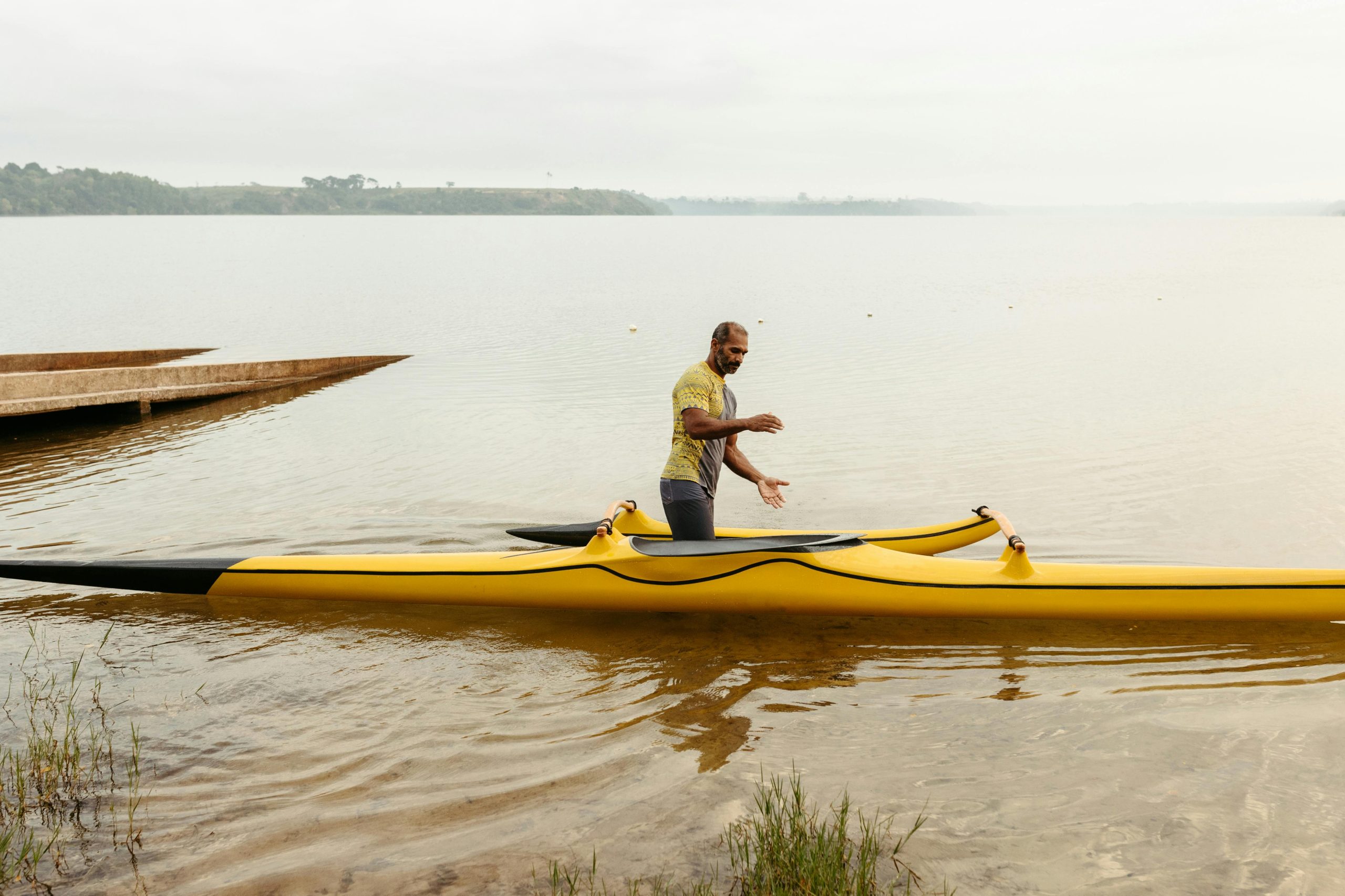 Man stands beside yellow kayak on a serene lakeshore, ready for outdoor adventure.