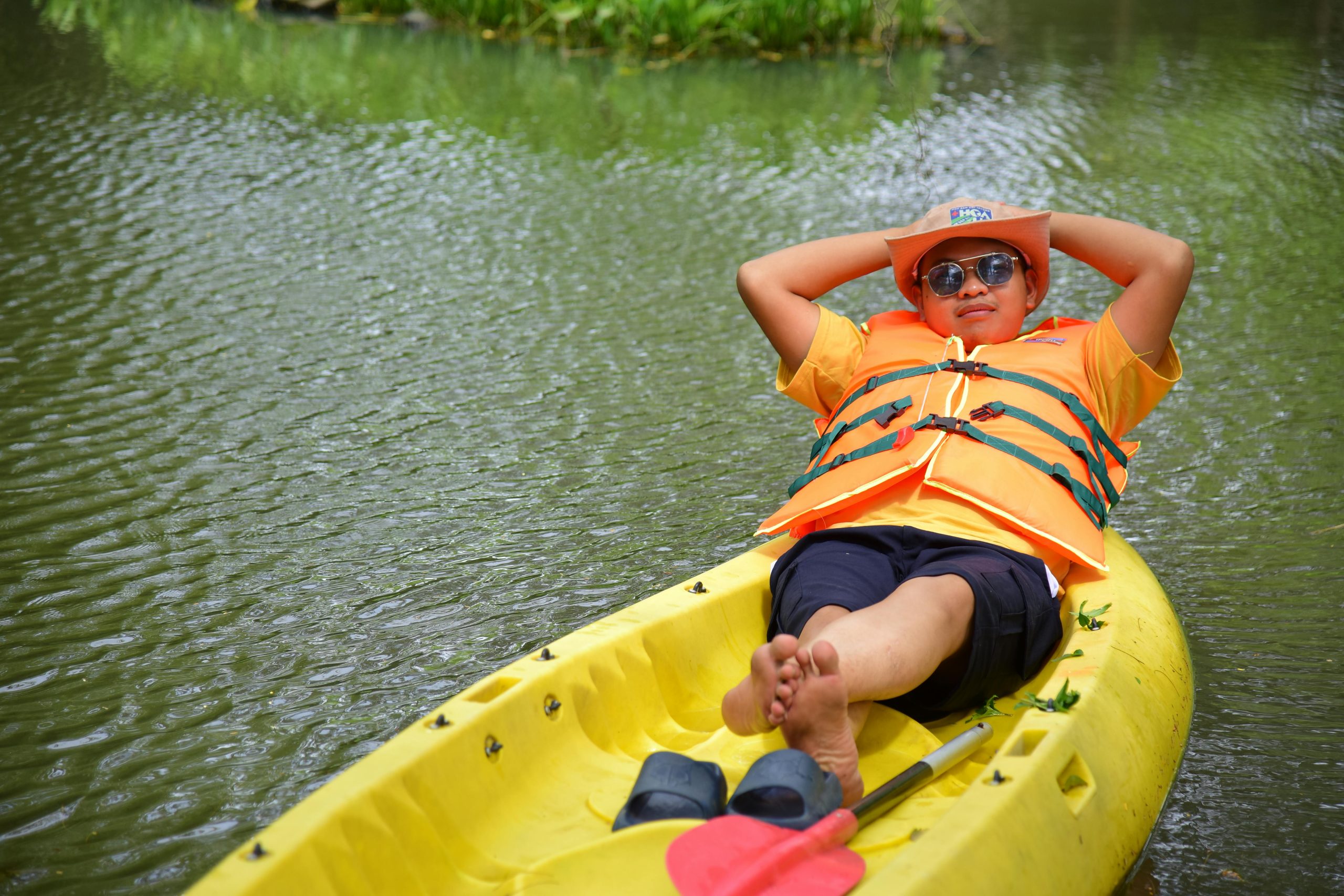 Man in orange life jacket relaxes on a yellow kayak with a hat and sunglasses on a calm lake.