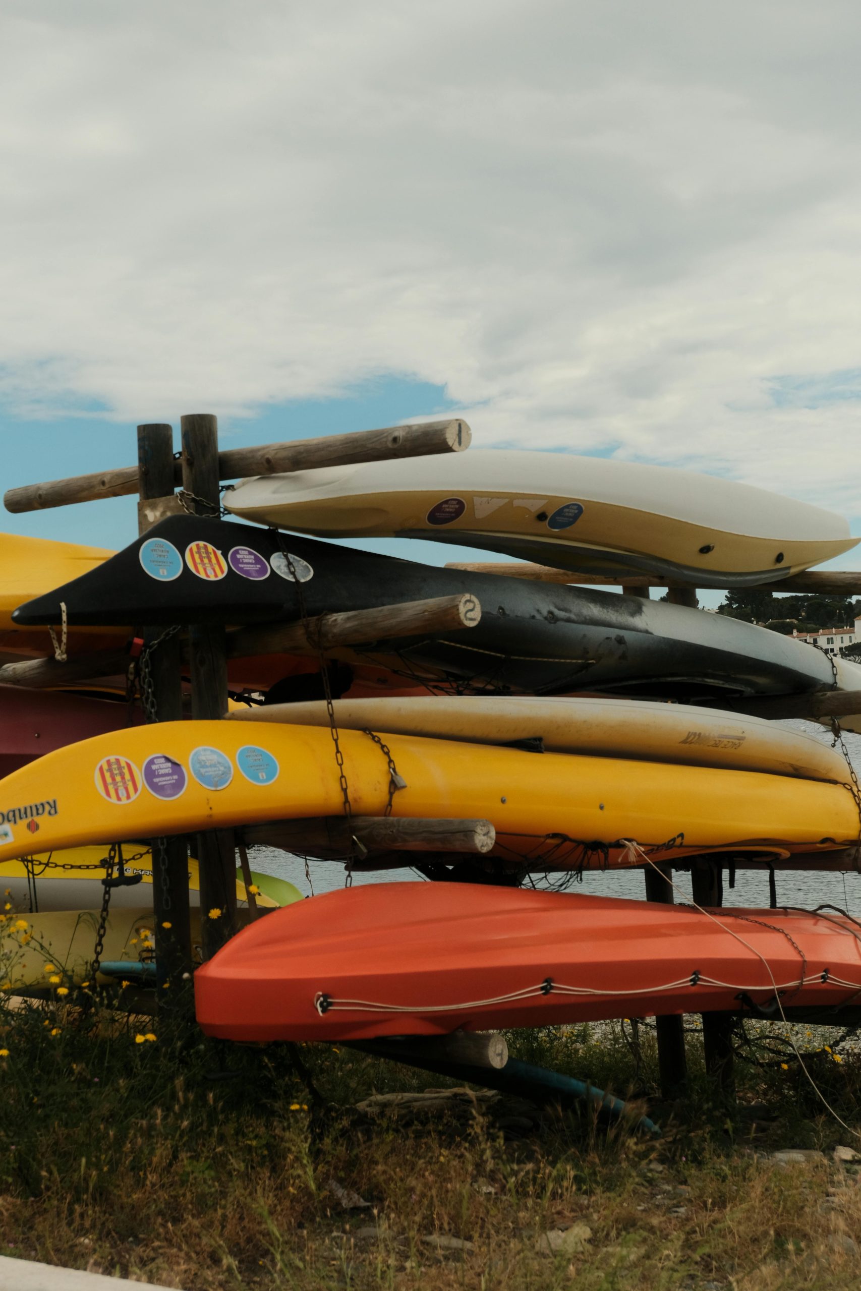Stack of colorful kayaks on a rack outdoors against a cloudy sky, suggesting adventure or leisure activities.