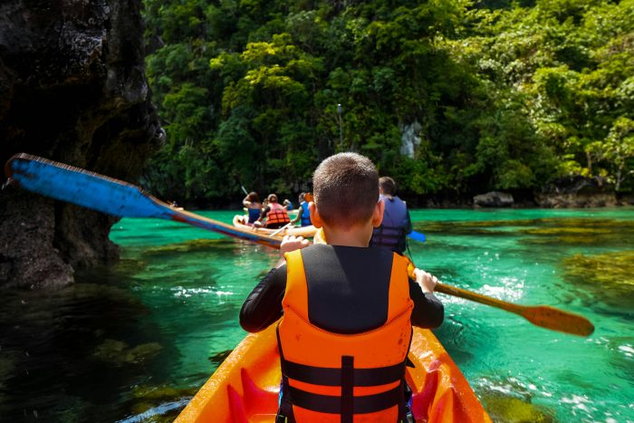 Group of children kayaking in turquoise water with lush green tropical forest in background.