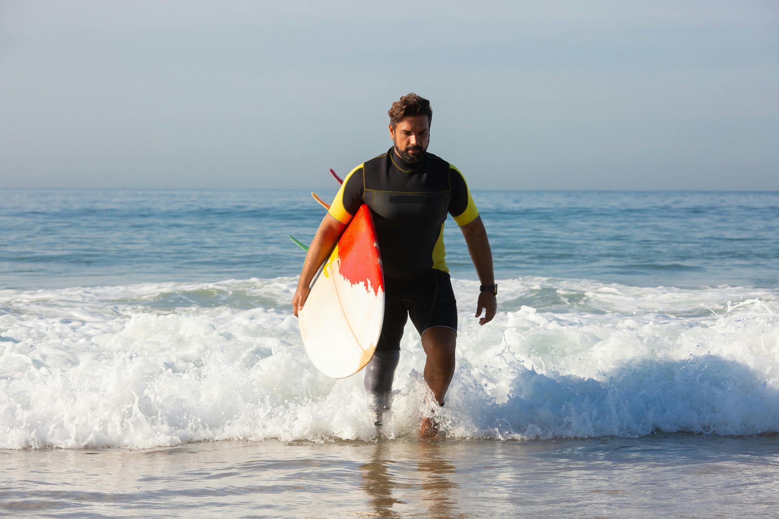 Full body sporty ethnic male surfer with leg prosthesis carrying surfboard and walking in foamy seawater towards sandy shore on sunny day