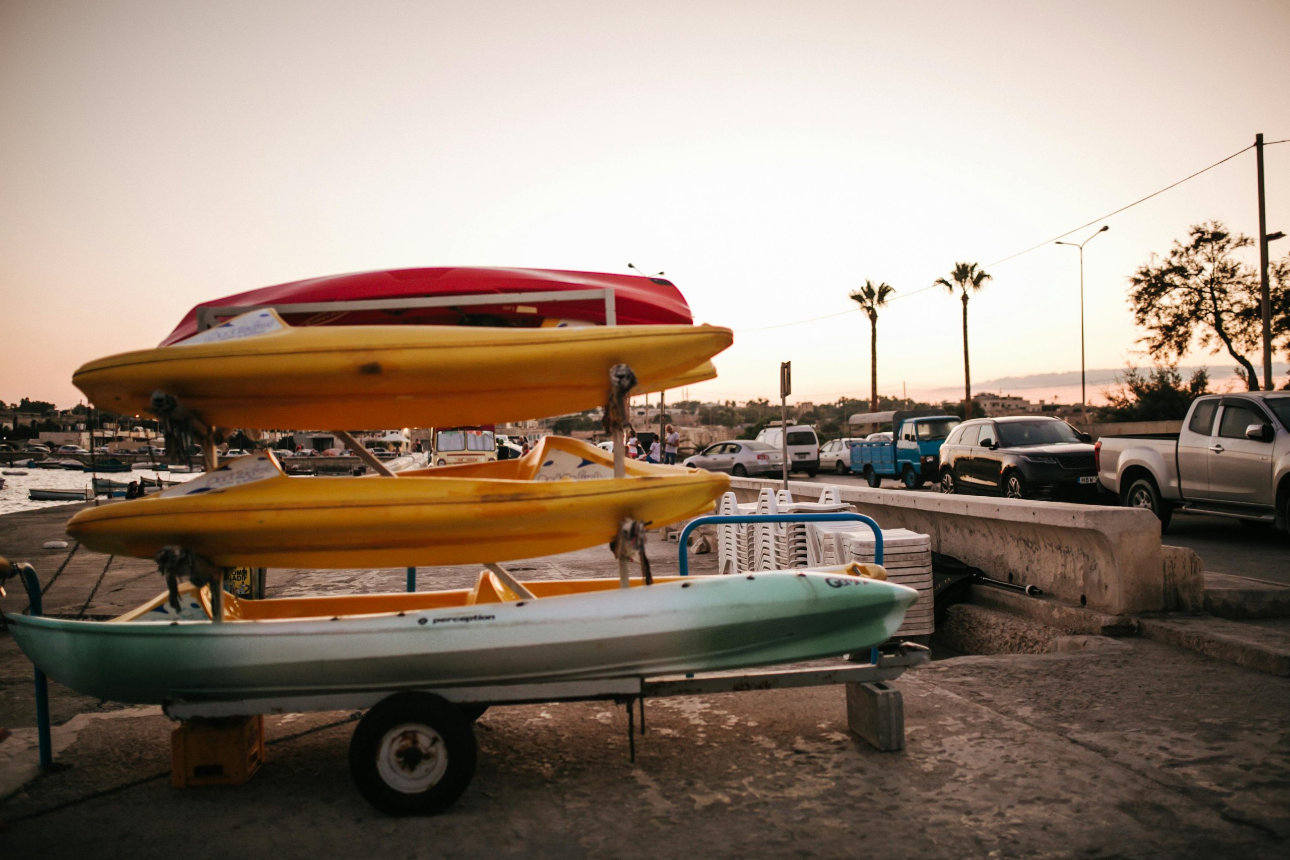 A stack of colorful kayaks at a marina during sunset, with parked cars nearby.
