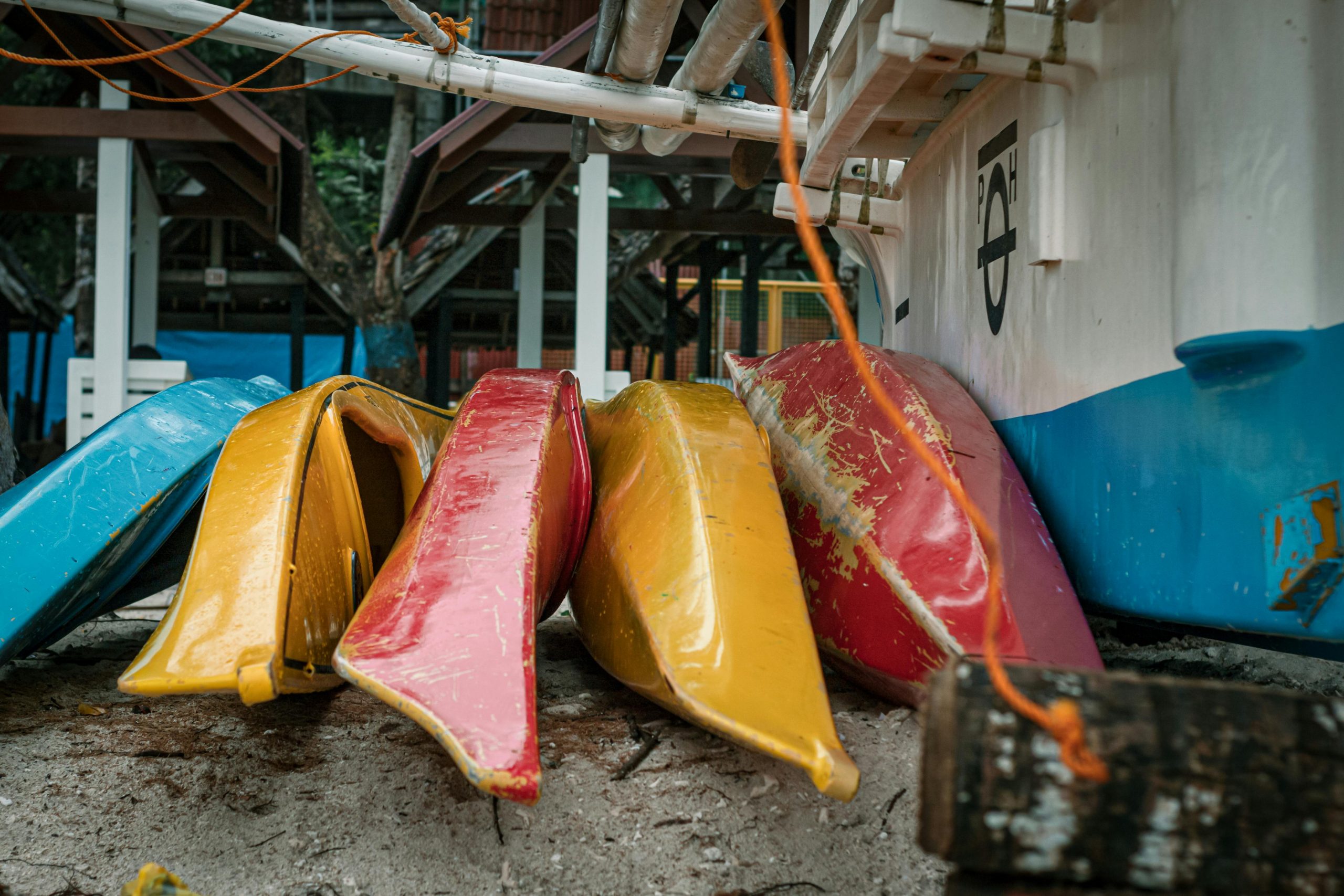 Vibrant yellow, red, and blue kayaks stacked on a sandy beach under a boathouse.