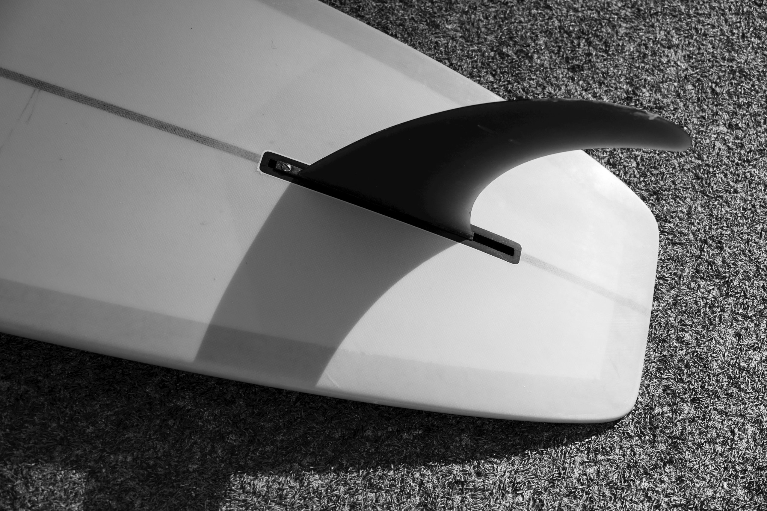 A black and white artistic capture of a surfboard with a shadowed fin on textured ground.