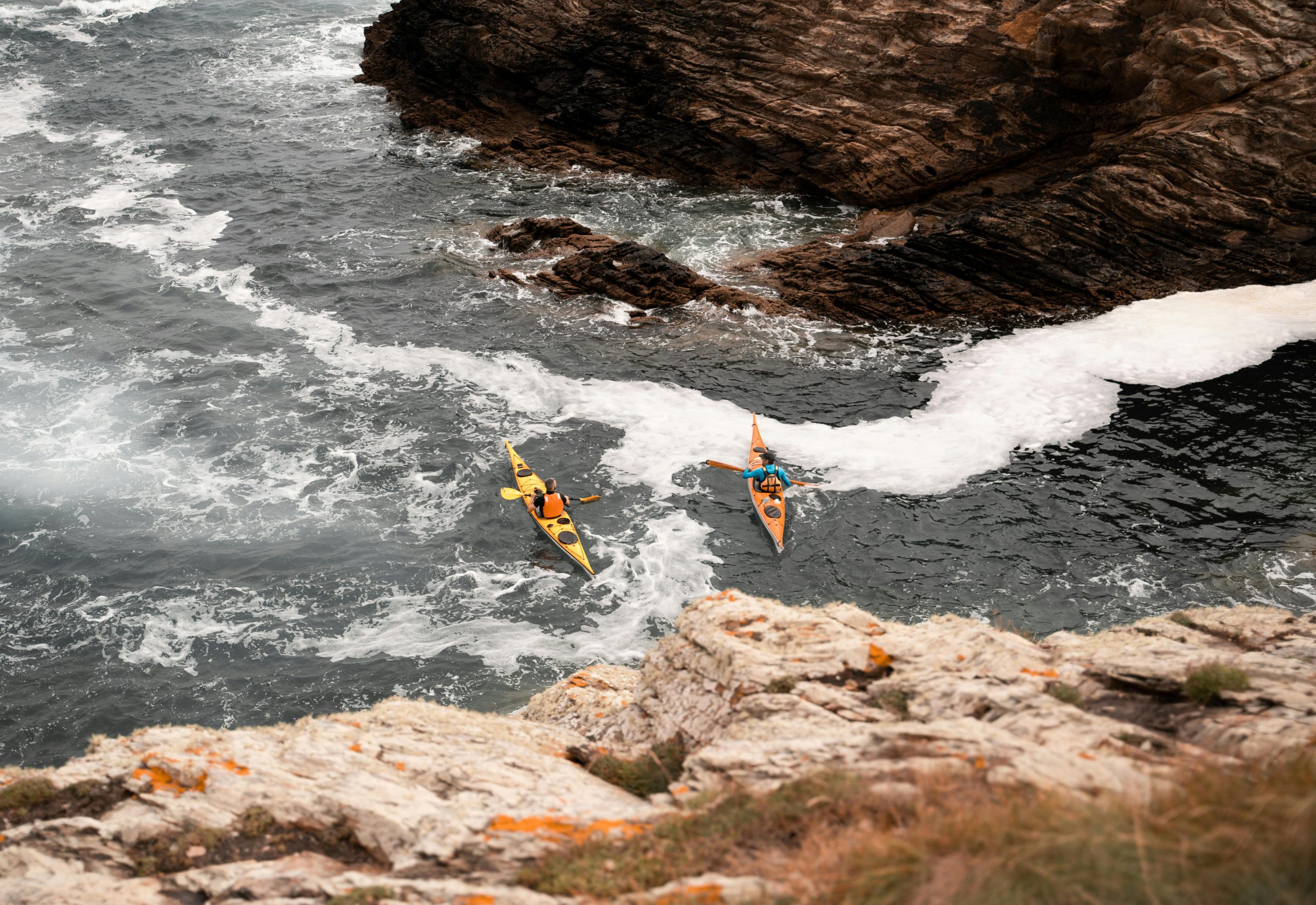 Two kayakers paddle through waves near rocky cliffs on a vibrant summer day.