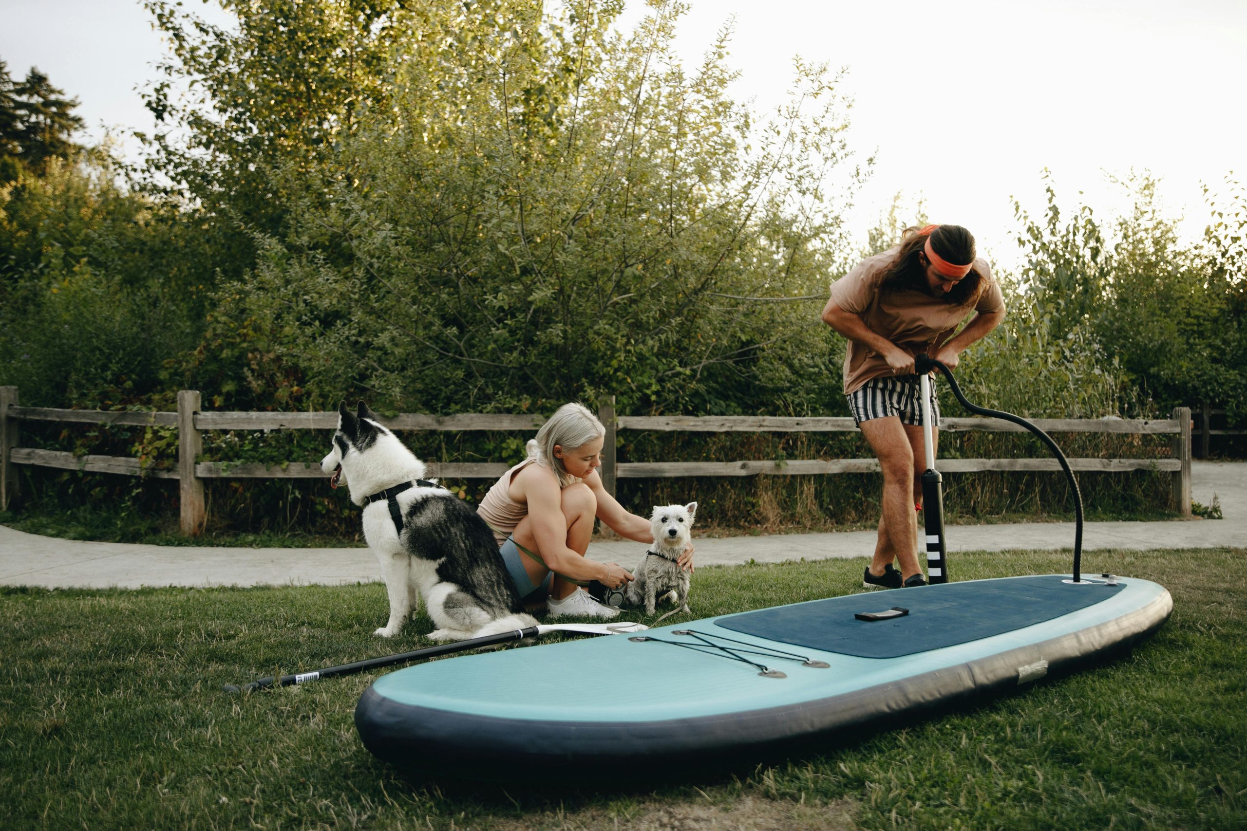 Man inflating paddle board while woman sits with dogs at park.