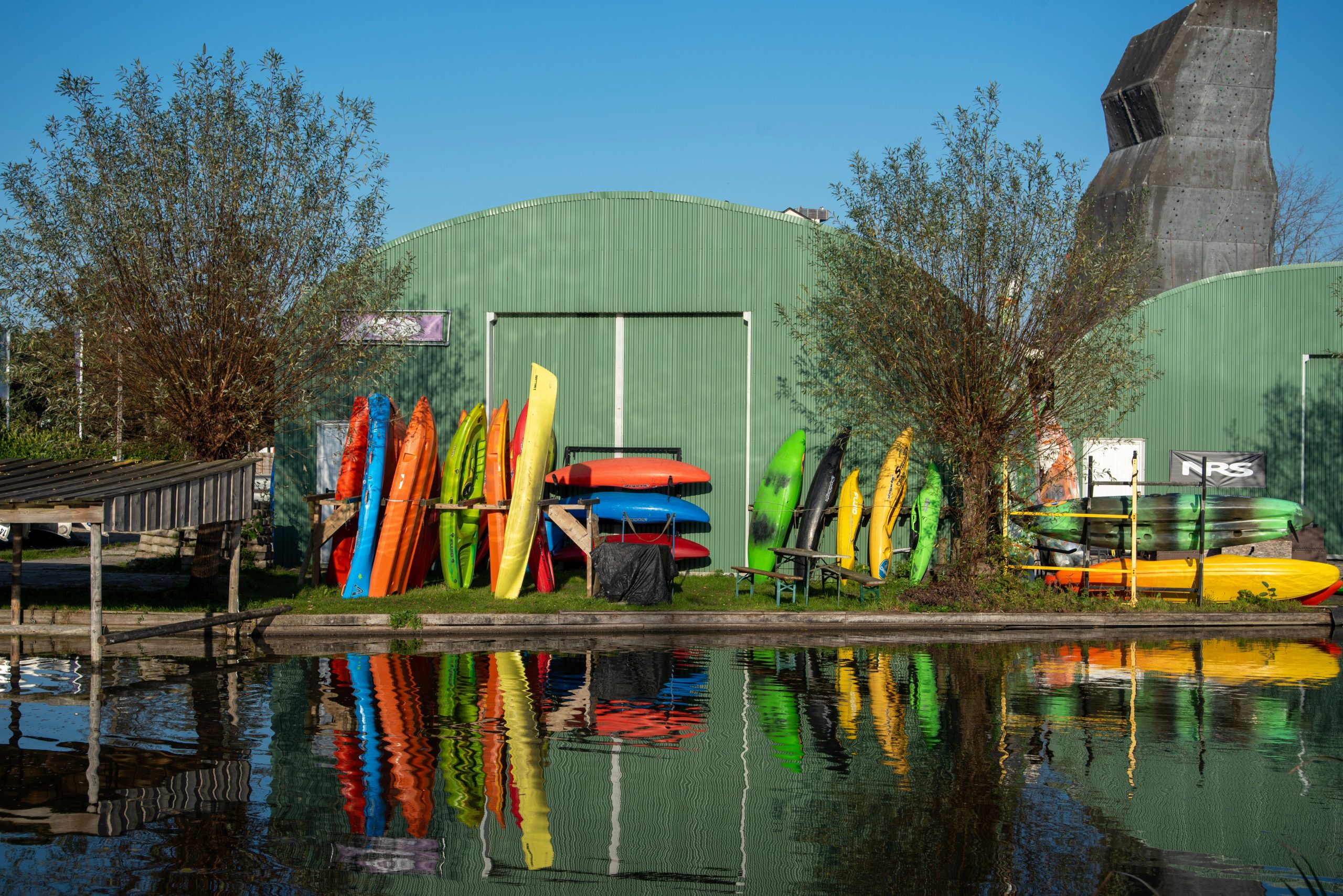 Vibrant canoes stacked by a green warehouse in Rotterdam, reflecting in water.