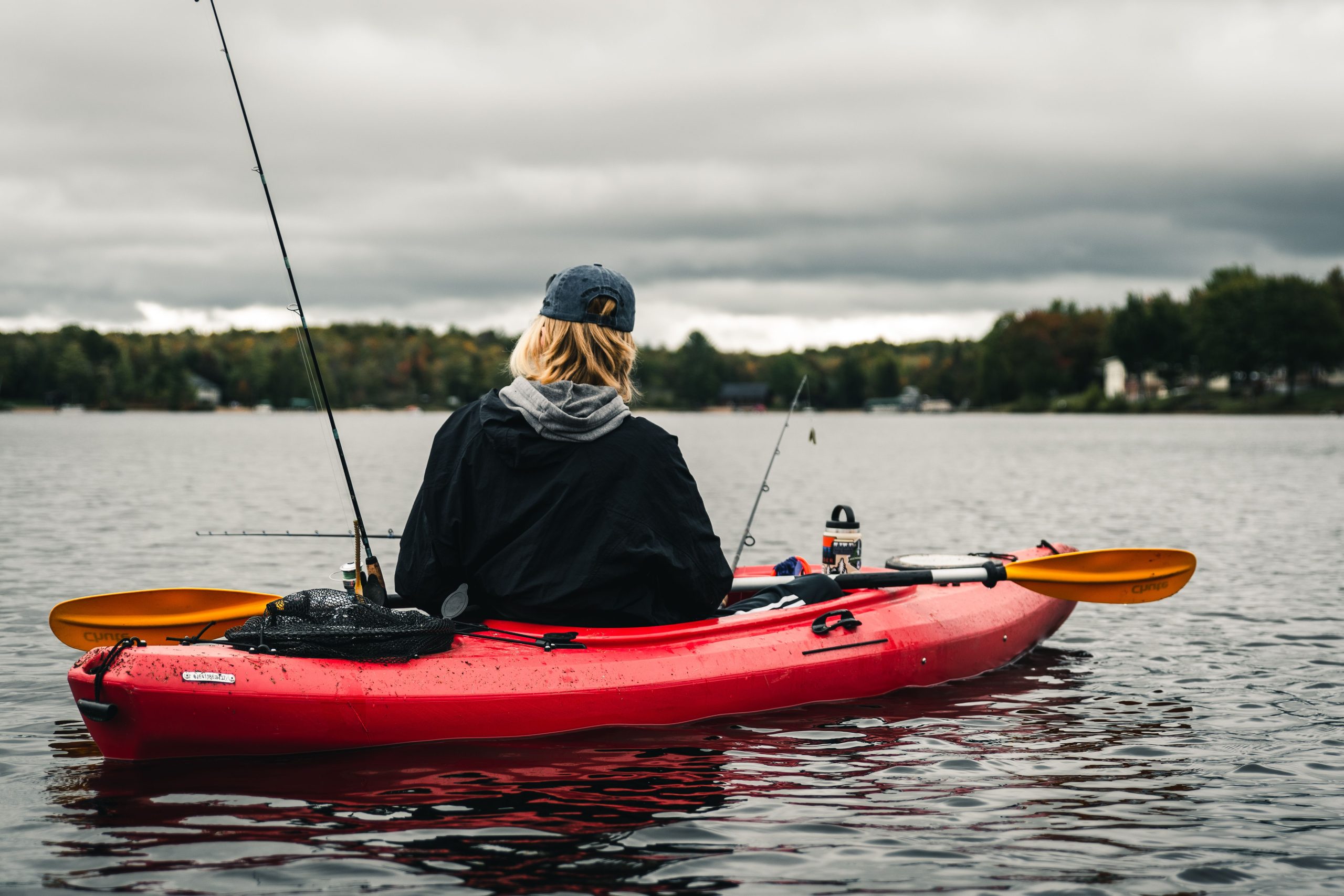 Person fishing in a kayak on a calm lake under cloudy skies, enjoying outdoor adventure.